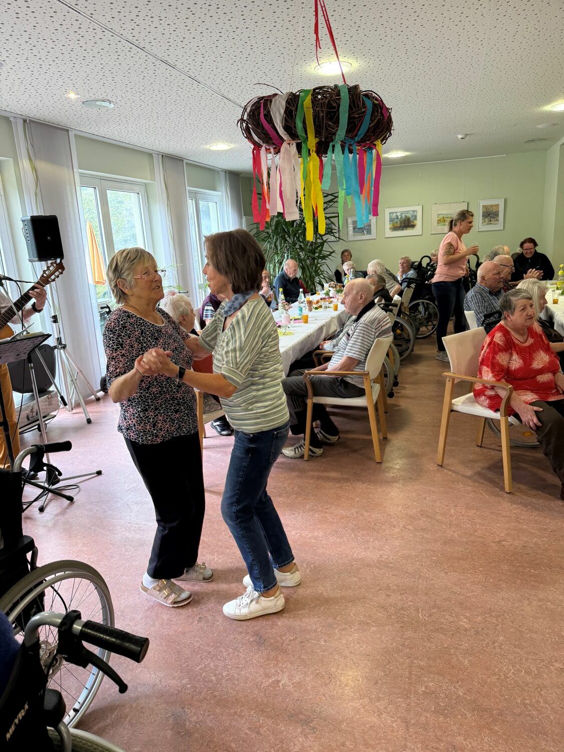 tanzende Seniorinnen und im Hintergrund die restlichen Bewohner*innen am Kaffeetisch
